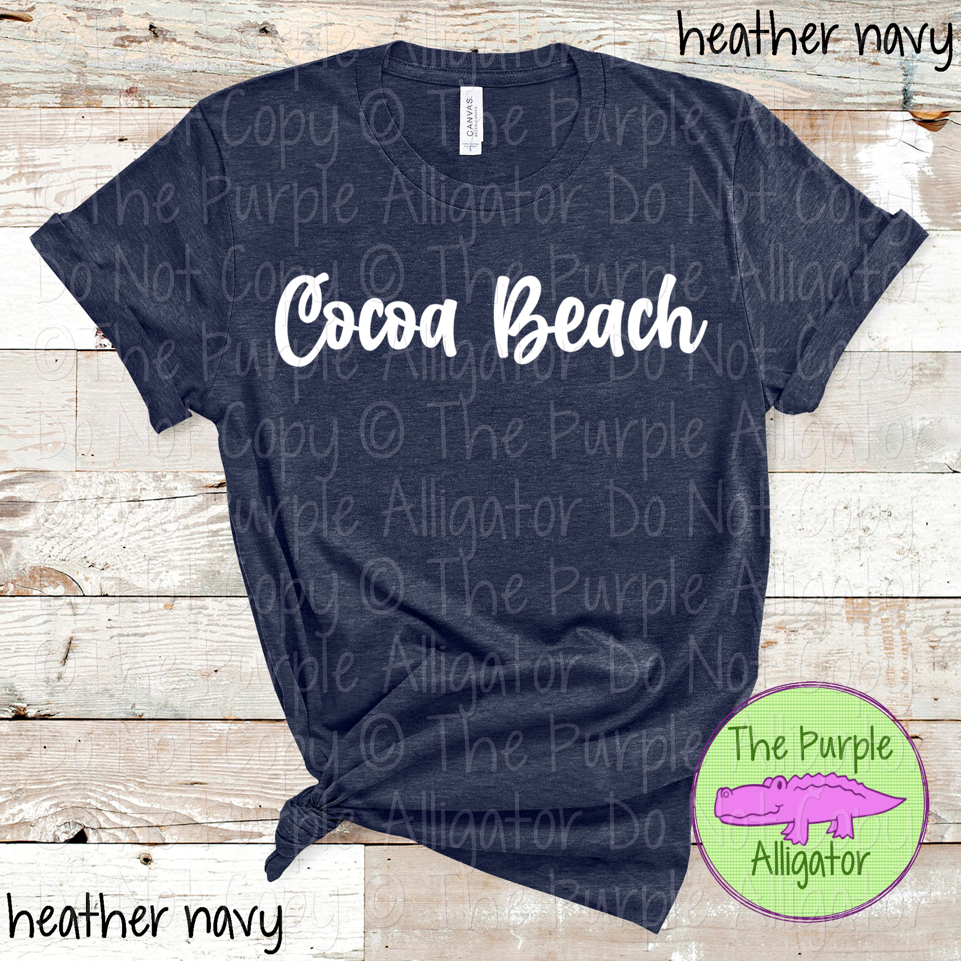 Navy t-shirt with 'Cocoa Beach' text on a wooden surface, branded with 'The Purple Alligator'.